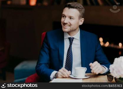 Joyful male worker in formal clothing looks happily aside, drinks hot aromatic coffee, happy to notice someone aside, sits in cafeteria, has pleasant smile on face. Cheerful businessman in restaurant