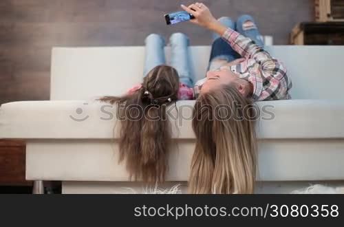 Joyful mother and cute daughter lying on sofa with long hair hanging down and making self portrait with smartphone as they spend free time together at home. Cheerful mom and little girl taking selfie on mobile phone in modern domestic interior.