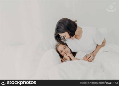 Joyful mother and daughter have fun together in bed, have happy expressions, enjoy comfort of soft bedclothes, have happy looks, spend morning in bedroom. Family, awakening and bedding concept