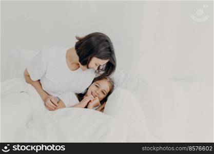 Joyful mother and daughter have fun together in bed, have happy expressions, enjoy comfort of soft bedclothes, have happy looks, spend morning in bedroom. Family, awakening and bedding concept