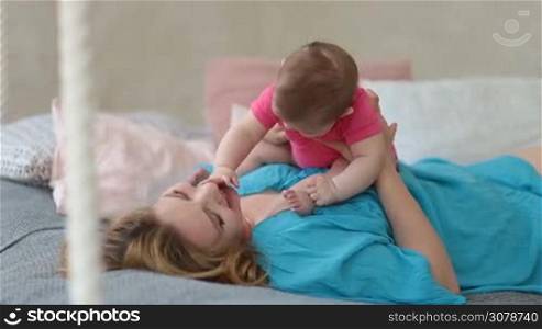 Joyful mother holding her baby girl and playing while lying on her back in bed in sunny bedroom. Young beautiful mom and infant daughter spending leisure at home, having fun, laughing