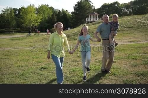 Joyful multi generation family holding hands dancing and jumping while walking across grass in park. Positive grandparents with grandchildren enjoying time, having fun and smiling on park lawn on sunny day. Slow motion. Steadicam stabilized shot.