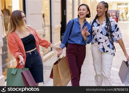 Joyful multiethnic female friends with shopping bags looking at each other with smile while meeting on street after shopping in city. Cheerful diverse women with shopping bags meeting on street