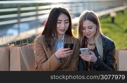 Joyful multiracial girlfriends watching media content online on mobile phone outdoors at sunset. Euphoric teenage girls after shopping watching video on smart phone and pointing at screen surprised while sitting on the bench in park.