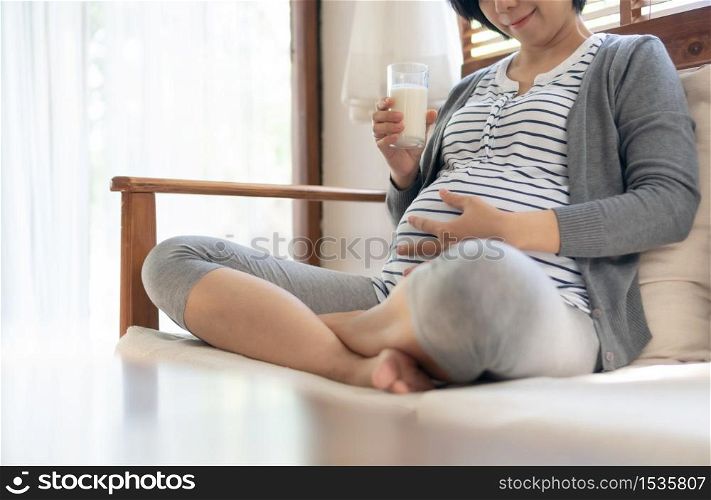 Joyful Pregnant young woman sitting drinking milk and touch her belly on sofa at home.