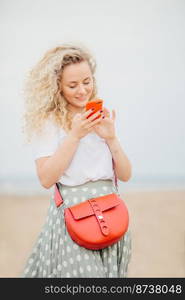 Joyful pretty young female with light curly hair, uses cell phone for surfing social networks, dressed in casual t shirt and skirt, recieves pleasant message from boyfriend, strolls at beach alone