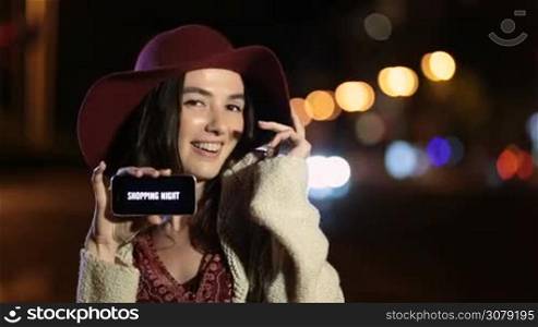 Joyful shopper woman in stylish hat showing smart phone with special offer - shopping night on black screen in night city street. Excited shopaholic female holding mobile phone with shopping night ad on screen at night over streetlights bokeh.