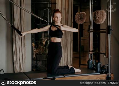 Joyful slender ginger woman on pilates reformer performing arm stretching exercise during training sessions in fitness studio, showing confidence in every move, sportive female wearing sports clothes. Fit slender woman on pilates reformer smiling during stretching exercises