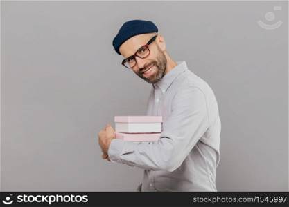 Joyful smiling bearded man with happy expression, dark bristle, wears white shirt and spectacles, carries presents, stands sideways at camera, models against grey background. Celebration concept
