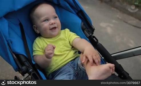 Joyful smiling cute infant child clapping hands in baby carriage while taking a stroll with father in summer park. Adorable toddler boy with blue eyes in pram enjoying leisure, happiness and joy during a walk outdoors. Slo mo. Stabilized shot.