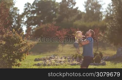 Joyful stylish father with beard throwing happy infant son high in the air in public park over colorful landscape background. Playful dad and cute toddler baby boy playing and having fun in nature while spending leisure together. Side view.