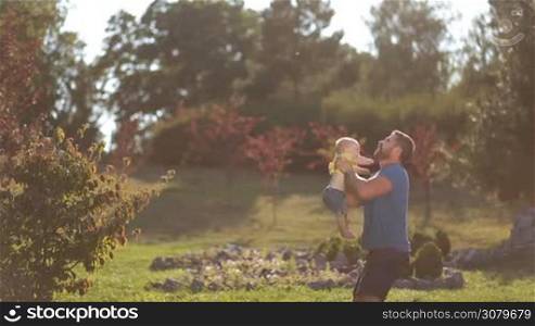 Joyful stylish father with beard throwing happy infant son high in the air in public park over colorful landscape background. Playful dad and cute toddler baby boy playing and having fun in nature while spending leisure together. Side view.