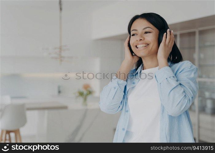 Joyful woman at home listens to music or podcasts with modern wireless headphones. Smiling, she enjoys her favorite songs and audio books with perfect sound quality.