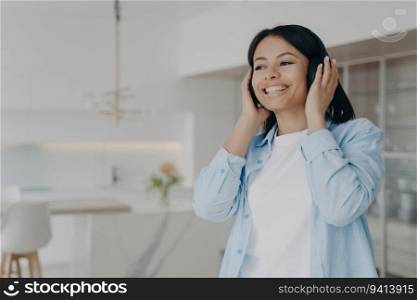 Joyful woman at home listens to music or podcasts with modern wireless headphones. Smiling, she enjoys her favorite songs and audio books with perfect sound quality.