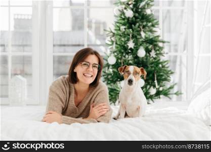 Joyful woman with short dark hair wears spectacles and round glasses, funny dog in spectacles poses near host, lie on bed, have good rest during Christmas eve, have fun together. New Year decoration