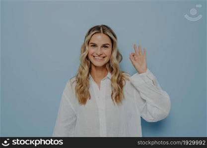 Joyful young blonde woman in white shirt making ok gesture with her hand in air, showing that everything is alright, signalling there is no problem while standing alone next to blue wall. Joyful blonde woman making ok gesture with hand
