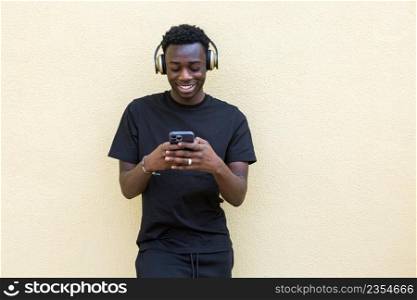 Joyful young confident African American guy in total black outfit and modern headphones listening to music and messaging on mobile phone standing near wall on street. Cheerful black male smiling and using smartphone while listening to music in headphones