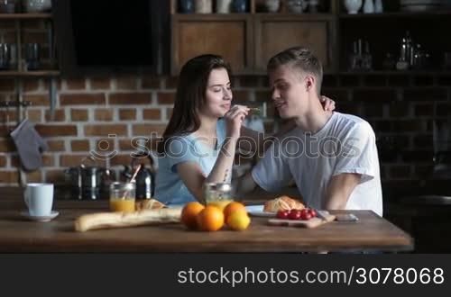 Joyful young couple in love sharing breakfast together while sitting at kitchen table in the morning. Pretty brunette girl feeding her handsome boyfriend and kissing. Smiling family spending great time and enjoying breakfast meal at home.