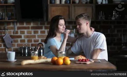 Joyful young couple in love sharing breakfast together while sitting at kitchen table in the morning. Pretty brunette girl feeding her handsome boyfriend and kissing. Smiling family spending great time and enjoying breakfast meal at home.