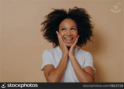 Joyful young dark skinned woman feeling happy, smiling at camera and touching cheeks with both hands, carefree african female in white tshirt expressing positivity, posing isolated on beige background. Joyful young dark skinned woman feeling happy, smiling at camera and touching cheeks with both hands