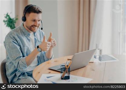 Joyful young guy looking at laptop screen and showing thumbs up while having online video call at home, looking at webcam and expressing positivity and agreement. Online communication concept. Happy bearded man during online call with friend at home