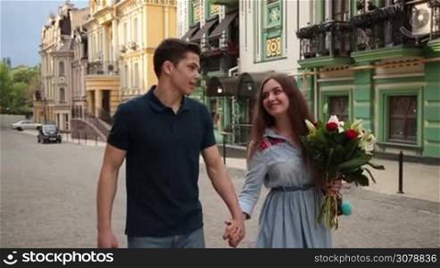 Joyful young hipster couple spending leisure together. Charming woman with bouquet of flowers holding hand of her handsome man walking along city street during romantic date. Slow motion. Steadicam stabilized shot.