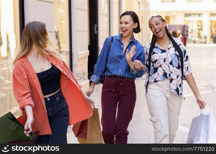 Joyful young multiracial female best friends in stylish clothes laughing and waving hair while walking on city street near fashion boutiques during shopping. Cheerful young diverse ladies having fun on street after shopping