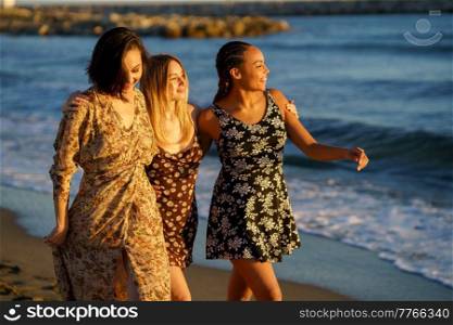 Joyful young multiracial female friends in sundresses walking together on sandy coast near waving sea while enjoying sunny summer day. Positive diverse women admiring seascape on beach