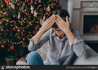 Joyful young woman in VR headset glasses sitting on cozy sofa next to decorated Xmas tree, amazed happy female in virtual reality googles closing eyes with hands, enjoying christmas holidays at home. Joyful young woman in VR headset glasses sitting on cozy sofa next to decorated Xmas tree at home