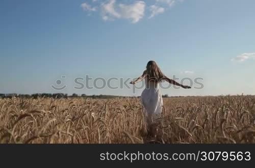 Joyful young woman in white dress with amazing long blond hair running through wheat field in summer against amazing skyline background. Positive beautiful female having fun and enjoying beauty of nature in countryside. Slo mo. Stabilized shot.
