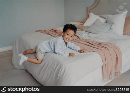 Joyous little mixed race boy kid laying and resting on his big comfortable bed with pillows, feeling happy and smiling while playing in bedroom. Children spending leisure time at home. Happy little afro american kid enjoying playtime at home