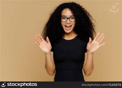 Joyous playful Afro American woman raises palms, feels excited, expresses happiness, glad to receive present, wears casual outfit, eyewear, stands against brown background. Positive reaction