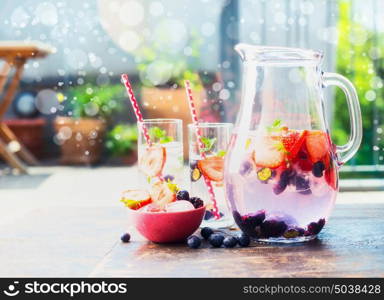 Jug and glasses with berries infused water on table in garden, front view