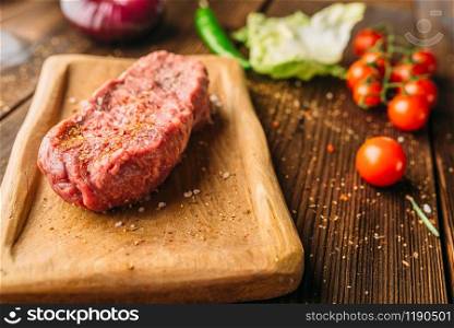 Juice piece of steak in seasoning and fresh vegetables on wooden table closeup, nobody. Uncooked beefstek, pepper and herb, food preparation, cooking. Juice piece of steak in seasoning and vegetables