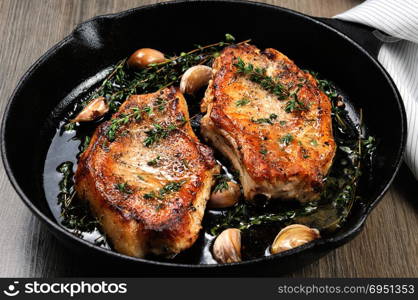 Juicy, browned pork steak on a bone in oil with garlic and herbs in a frying pan. Close-up.