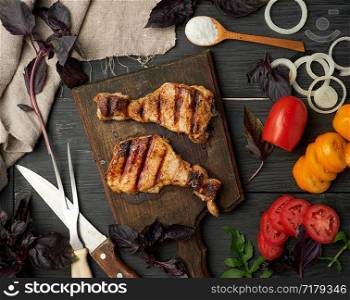 juicy grilled pork steaks on a brown wooden board, fresh red and yellow tomatoes