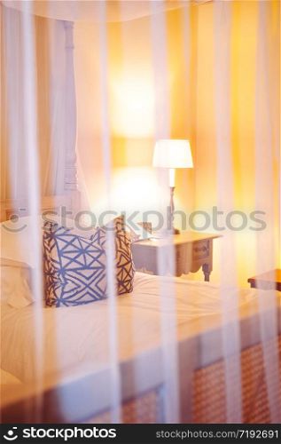 JUN 18, 2011 Serengeti, Tanzania - African asian contemporary hotel bedroom with wooden four poster bed, pillow and table lamp seen through white curtain. under warm tone light
