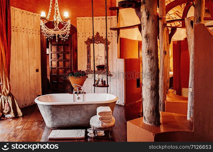 JUN 23, 2011 Ngorongoro, Tanzania - Luxury African tribal hut bathroom interior decoration with old vintage ceramic bathtub and chandelier with colonial furniture and glamourous ornaments under warm light