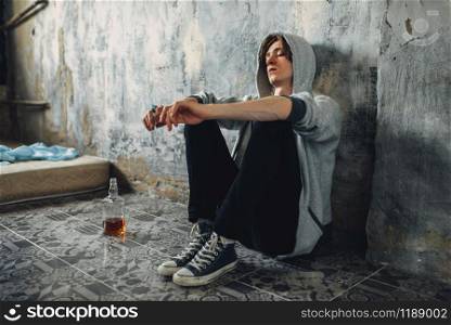 Junkie with syringe in hand sitting on the floor after dose, bottle of alcohol is near. Drug addiction concept, narcotic addicted people. Junkie with syringe sitting on floor after dose