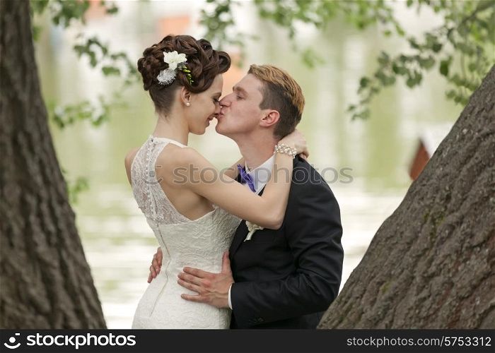 Just married couple kissing between tree trunks and pond&#xA;