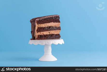 Just one piece of chocolate birthday cake with cocoa frosting, on a white stand and blue background. Minimal piece of cake. Festive layered cake slice