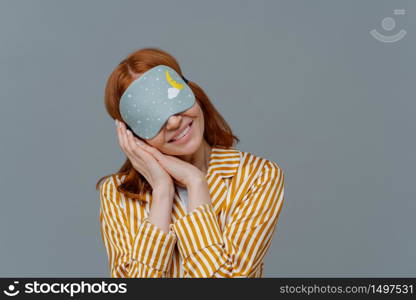 Just relaxing or time for nap. Smiling gentle woman with eye mask, tilts head on pressed palms, wears striped pajama, poses over grey wall, copy space area for your text feels comfortable and relaxed