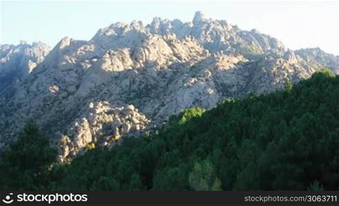 Kahler steiniger wei?er tropfsteinformiger Berg hinter grunen Walder aus dicht bewachsenen Tannen und Laubbaumen. Die Sonne strahlt auf den Berg.