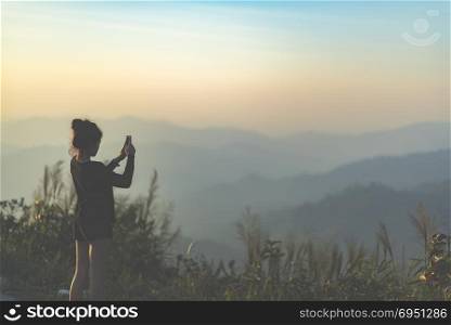 Kanchanaburi, Thailand - March 14, 2016: Woman is taking a photo of a beautiful sunset above the mountain with her mobile smart phone, vintage filtered Images