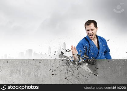 Karate man in blue kimino. Young determined karate man breaking with hand concrete wall