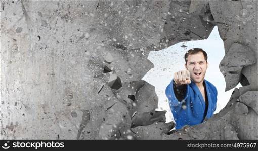 Karate man in blue kimino. Young determined karate man breaking with hand concrete wall