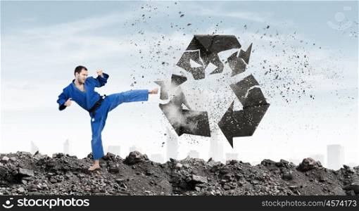 Karate man in blue kimino. Young determined karate man breaking with leg concrete recycle sign