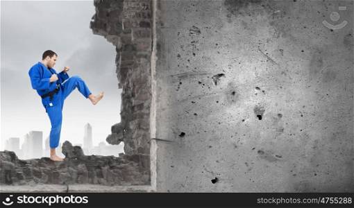 Karate man in blue kimino. Young determined karate man breaking with leg concrete wall
