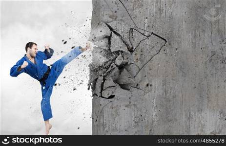Karate man in blue kimino. Young determined karate man breaking with leg concrete wall