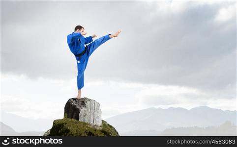 Karate man in blue kimino. Young determined karate man on rock top high in sky